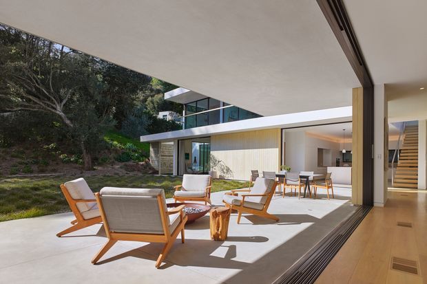 A bright contemporary indoor-outdoor living space opening onto a landscaped garden. The covered terrace features timber-framed lounge chairs, a low table and an outdoor dining setting, while large retractable glazed doors connect it to a minimalist interior with a kitchen, dining area and staircase. The building has strong horizontal lines, flat roof planes, extensive glazing and pale rendered surfaces.