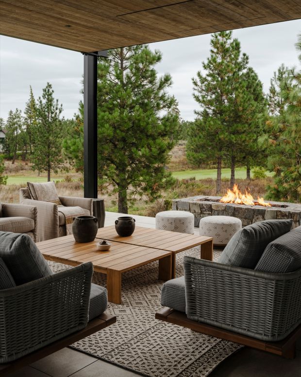 Covered outdoor terrace furnished as a relaxed lounge, with woven armchairs and upholstered seating arranged around a timber coffee table. A stone outdoor fireplace with a live fire sits beyond the seating, overlooking a landscaped pine setting. The terrace has a warm timber ceiling, dark structural post, and patterned area rug.