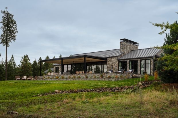 A low, contemporary single-storey house set in a landscaped rural or wooded site. The building features a dark standing-seam metal gabled roof, extensive floor-to-ceiling glazing, a broad covered outdoor terrace with slim dark posts, and a prominent rough natural-stone chimney and wall elements. Stone retaining walls and native-looking planting frame the lawn and outdoor seating area.