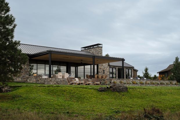 A low, contemporary stone-clad house set in a landscaped grassy site. The building features extensive floor-to-ceiling glazing, slender dark metal framing, broad covered outdoor terraces, a low-pitched standing-seam metal roof, and a prominent stone chimney or masonry volume. Outdoor seating and steps connect the house to the lawn.