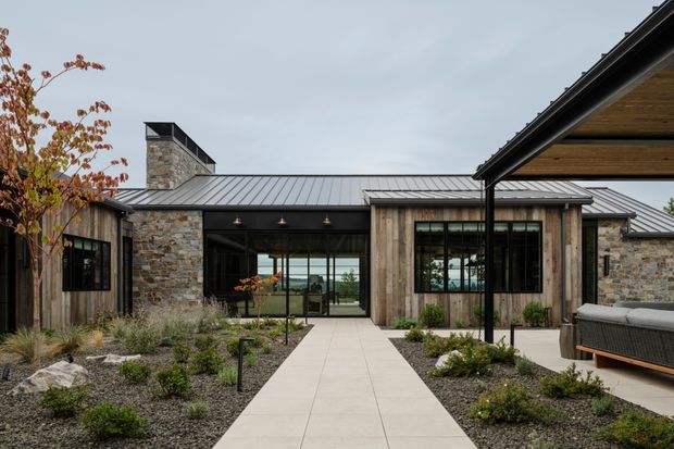 A low, modern single-storey residence viewed from a landscaped courtyard. The building combines dark standing-seam metal pitched roofs, natural stone masonry, vertical weathered timber cladding, and extensive dark-framed glazing. A broad pale paved path leads to a recessed glazed entry, with outdoor seating and a covered terrace visible at right. Native-style planting, gravel beds, and autumn foliage frame the approach.