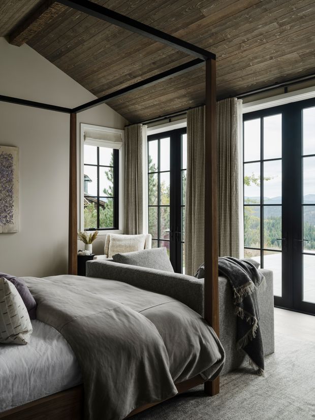 A refined bedroom with a partially visible timber bed in the foreground, a grey upholstered lounge chair, neutral bedding, and full-height black-framed glazed doors opening toward a scenic landscape. The room features a dark exposed timber ceiling with black structural beams, warm timber posts, soft woven curtains, pale walls, and a large neutral rug. The overall aesthetic is calm, contemporary, and natural with strong indoor–outdoor connection.