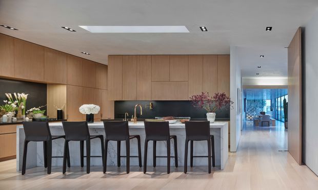 A refined open-plan kitchen with a long central island and six dark upholstered bar stools. The kitchen features extensive warm timber cabinetry, a dark stone or composite splashback, pale stone benchtops, integrated appliances, and a large skylight. A bright hallway connects to a living area visible in the distance, with pale timber flooring and restrained contemporary detailing throughout.