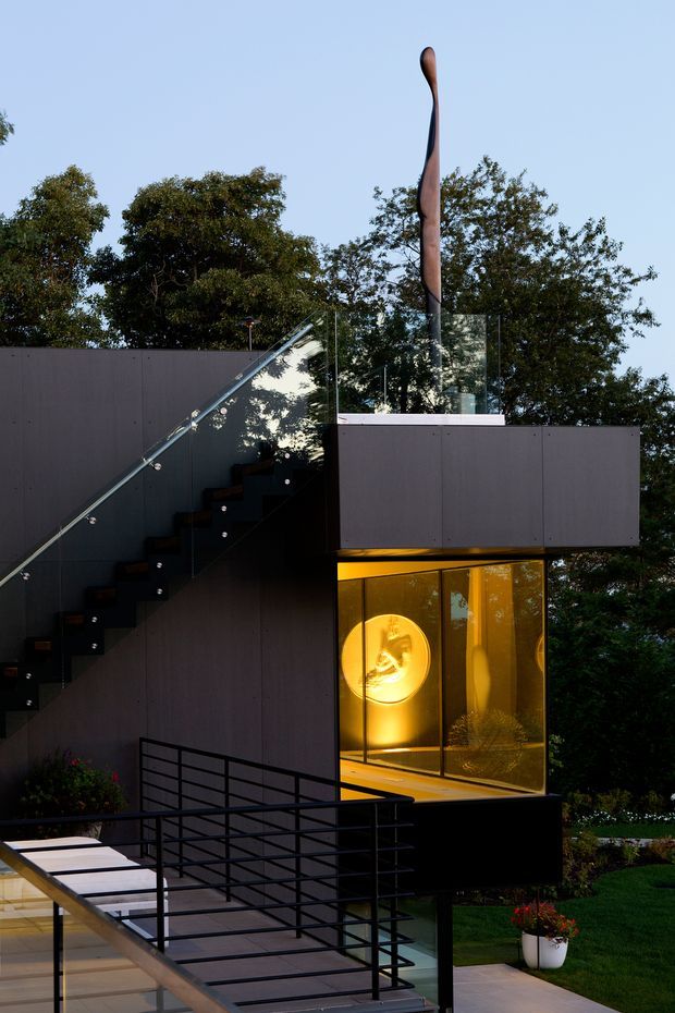 Exterior view of a contemporary multi-level house set among mature trees, featuring dark smooth cladding, projecting box-like volumes, extensive clear glazing, and an exterior stair with glass balustrade. Warm amber interior lighting glows through the lower-level window, while a tall sculptural element rises above the roof terrace. Black metal railings, paved terraces, and landscaped lawn are visible in the foreground.