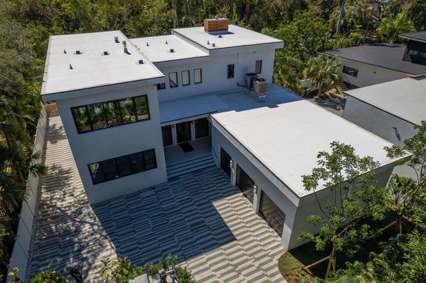 Aerial view of a contemporary multi-level residence with crisp white rendered walls, several low-pitched flat roofs, dark-framed windows, and a large patterned tiled courtyard or terrace connecting the building wings. Dense mature vegetation surrounds the property, with neighboring modern homes visible in the background.