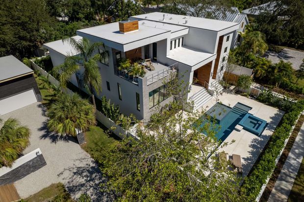 Aerial view of a contemporary multi-storey residence with crisp white rendered walls, flat roof forms, dark-framed glazing, and warm timber-clad accents. The home includes multiple terraces and balconies, landscaped gardens, paved outdoor areas, and a rectangular swimming pool with an integrated spa or shallow section. A separate flat-roofed garage is visible at the left.