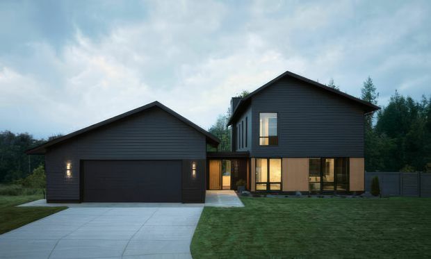 A contemporary two-part detached home photographed at dusk, with dark horizontal cladding, steep gabled roofs, warm timber-toned lower-level accents, and large illuminated windows. A broad concrete driveway leads to an attached double garage, with a manicured lawn and wooded backdrop.