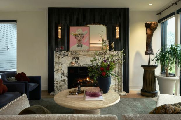 Sophisticated living room with a marble fireplace framed by a dramatic black fluted feature wall, an arched mirror, artwork, sculptural decorative objects, and layered furnishings. A round light timber coffee table sits centrally, with dark blue seating, a large rug, indoor plants, and expansive windows creating a refined, eclectic atmosphere.
