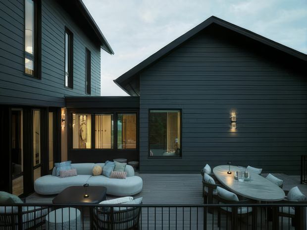 An evening view of a contemporary multi-level home with dark horizontal cladding and steep gabled roof forms, enclosing a timber deck courtyard. The deck is furnished as an outdoor living and dining area, with a curved pale sofa, coffee table, dining table, outdoor chairs, and subtle integrated lighting. Black balustrading frames the foreground, while glazed doors and windows connect the deck to the interior.