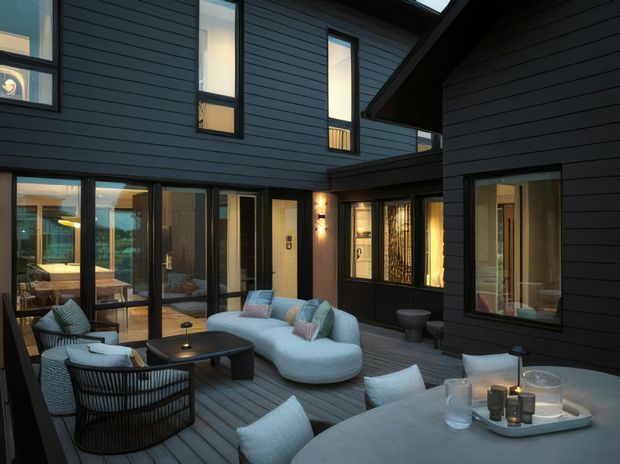 A sophisticated enclosed courtyard terrace between dark-clad building volumes, furnished as an outdoor lounge. The space features charcoal horizontal cladding, extensive floor-to-ceiling glazing, a timber deck, a curved light-colored sofa, woven lounge chairs, low dark tables, and warm wall lighting. Interior spaces are visible through the surrounding glass doors and windows.