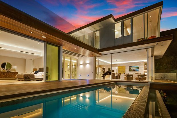 Twilight exterior view of a sophisticated multi-storey contemporary residence arranged around a courtyard-style swimming pool. Extensive floor-to-ceiling glazing opens the living spaces to timber decking and the pool, while projecting upper-level volumes, clean rooflines, recessed lighting, and warm timber soffit cladding define the architecture. A bedroom and open-plan living area are visible through the glazing.