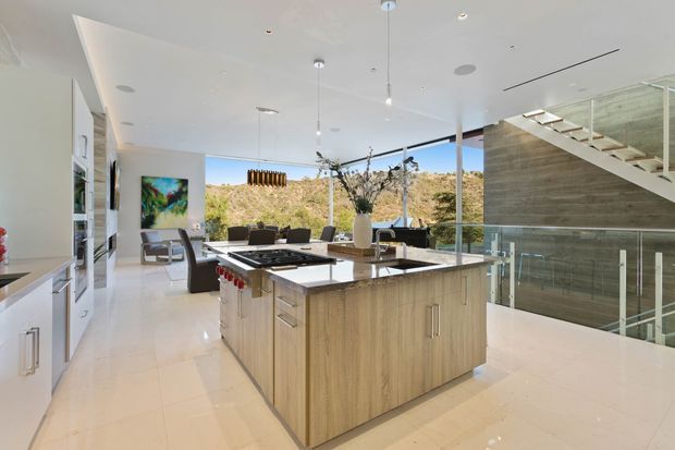 A bright, open-plan contemporary kitchen with a large light-wood island, pale stone benchtops, white built-in cabinetry and glossy pale flooring. The island includes integrated cooking and seating zones, while a dining/living area opens toward expansive glazing and a landscaped hillside view. A glass balustrade and open stairway are visible on the right, contributing to the spacious, high-end interior.
