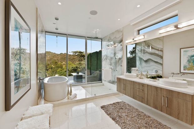 A bright, spacious contemporary bathroom with a freestanding oval bathtub beside floor-to-ceiling glazing, a glass-enclosed shower, double vanity with timber-look cabinetry, large illuminated mirror, and dramatic veined stone wall surfaces. Polished pale flooring and a neutral palette create an airy, luxurious interior overlooking greenery.