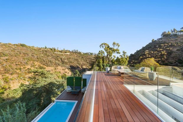 A sunny elevated outdoor terrace with a long reddish timber deck overlooking a steep, dry landscaped valley. A narrow rectangular swimming pool sits beside the deck, while clear glass balustrades protect the terrace and stair access. The setting is contemporary and open, with sculptural outdoor seating and planters, surrounded by rugged hillside vegetation under a clear blue sky.