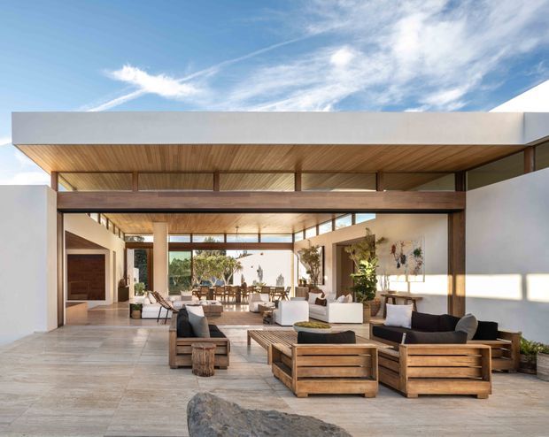 A spacious, open-air contemporary living pavilion with a broad flat roof and deep timber-lined soffit. White rendered walls frame expansive openings with dark timber or metal structural beams and clerestory glazing. The relaxed seating area is furnished with low timber-framed sofas and armchairs on a large pale stone or concrete-tile floor, opening toward a landscaped courtyard and dining area. The composition is bright, warm, and indoor-outdoor in character.