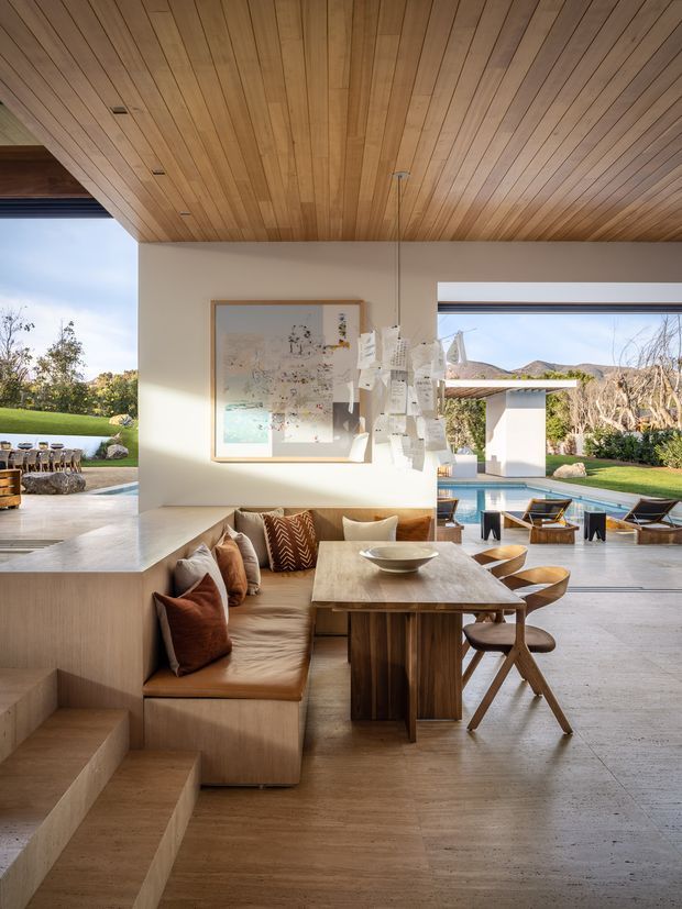 Open-plan dining and living area with a built-in upholstered bench, timber dining table and chairs, and a sculptural paper pendant light. Warm timber slat ceilings, pale walls, and stone flooring frame large openings to an outdoor pool terrace and landscaped garden. A short set of steps connects the dining area to a lower seating zone, creating a relaxed indoor-outdoor setting.