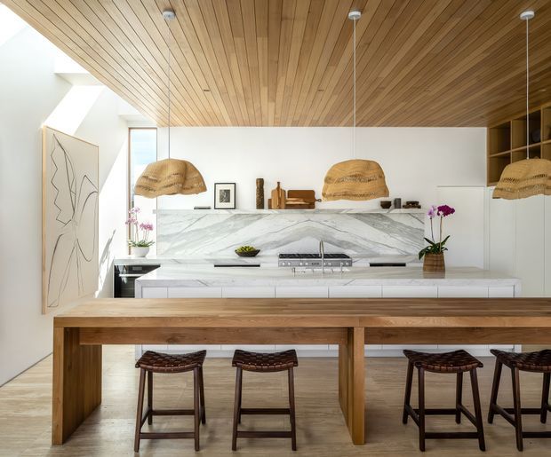 Bright contemporary kitchen and dining area with a long solid timber island or dining bench, dark woven bar stools, white cabinetry, a dramatic veined stone splashback, timber-lined ceiling, open shelving, sculptural woven pendant lights, and a skylit or clerestory-lit end wall. The space has a warm, refined natural-material palette and an open-plan arrangement.