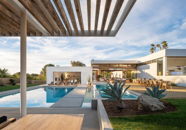 A bright contemporary residential outdoor entertaining area viewed from beneath a timber pergola. The scene features a large swimming pool with a central paved walkway, landscaped raised garden beds with sculptural agave plants and rocks, a white flat-roofed house with extensive glazing, and a covered dining and lounge terrace. Timber pergola beams, concrete paving, warm timber furniture, and blue sky create an open, resort-like setting.