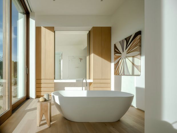 Bright contemporary bathroom with a freestanding white oval bathtub positioned in front of full-height warm timber cabinetry and a large mirror. Floor-to-ceiling glazing along the left wall brings in strong natural light, while pale walls, timber flooring, minimalist fittings, and abstract wall art create a refined, airy atmosphere.