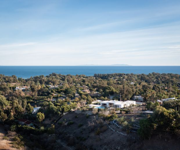 Wide elevated exterior view over a leafy coastal residential landscape, with a modern low-rise white building in the foreground, dense vegetation and scattered homes, and the blue ocean with a distant low island or headland on the horizon. The image is an architectural site and setting photograph dominated by the coastal outlook.