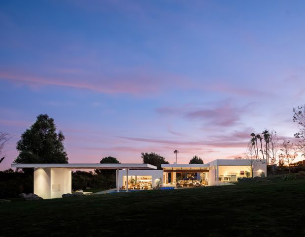 A low-slung modern residence viewed from the landscaped grounds at dusk. The house has a long, horizontally composed form with flat roofs, extensive floor-to-ceiling glazing, illuminated open-plan interiors, and a projecting covered outdoor structure. Dark lawn and mature trees frame the building against a pink and blue evening sky.