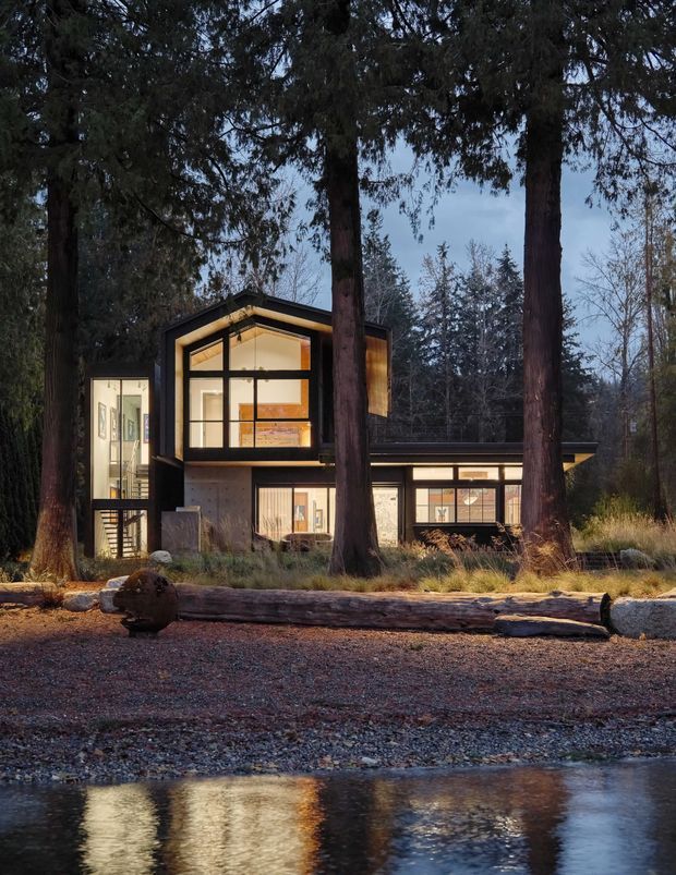Exterior view of a contemporary multi-level house nestled among tall trees beside a calm body of water. The home features dark structural framing, extensive warm-lit glazing, a prominent asymmetrical gable-like roof volume, and lower horizontal wings. Natural timber and concrete-toned surfaces blend with the forest setting, while a gravel shoreline and fallen log occupy the foreground.