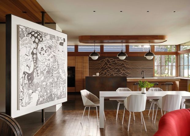A sophisticated open-plan dining and kitchen interior with warm timber cabinetry, polished dark wood flooring, and expansive windows. A white dining table with sculptural faceted white chairs sits beneath a long timber pendant feature with three spherical lights. The kitchen includes a dark island, timber joinery, and a dramatic dark textured feature wall, while a large monochrome artwork and red curved seating add visual contrast.