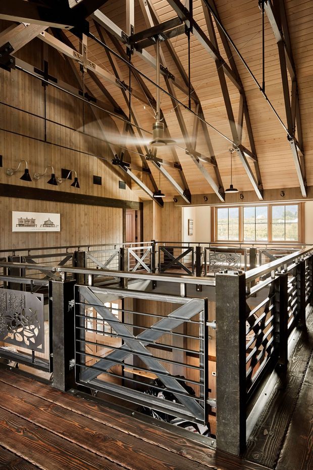 Interior view of a spacious timber-lined multi-level space with a high pitched ceiling and exposed dark metal and timber trusses. The image is taken from an upper-level landing or mezzanine overlooking a lower floor, with industrial-style metal railings, cable detailing, pendant lights, and a large ceiling fan. Warm wood surfaces contrast with black structural elements, while large windows bring in natural light.