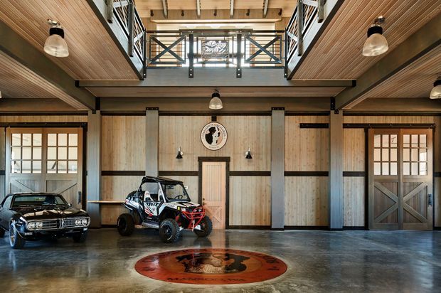 A spacious, rustic-industrial garage or workshop with polished concrete flooring, timber-lined walls and ceilings, exposed dark structural beams, and a double-height mezzanine with black metal railings. Large timber-framed doors flank the room. A classic car and off-road buggy are displayed on the floor, with pendant and wall-mounted industrial lighting and a circular floor emblem adding visual interest.