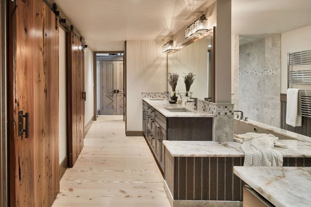 A spacious, refined bathroom featuring a long double vanity with dark timber cabinetry, pale marble or stone benchtops, patterned tile splashback, large mirror, and wall-mounted lighting. A freestanding or partially visible bathtub occupies the foreground, while a tiled shower area and heated towel rail are visible to the right. The room connects to a corridor lined with tall timber doors and pale timber flooring.