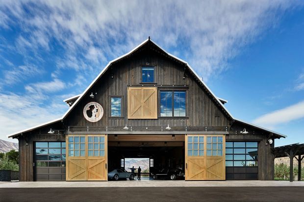 Front exterior view of a large barn-inspired building with dark vertical timber cladding, a steep gabled roof, and prominent timber-framed garage or barn doors. The open central bay reveals vehicles and a distant landscape, while glazed side doors and upper windows add natural light. A covered timber structure extends from the right side. The image is photographed in bright daylight beneath a blue, lightly clouded sky.