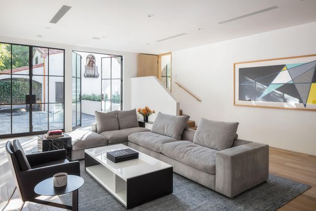 Bright contemporary living room with a large grey sectional sofa, black-and-white coffee table, lounge chair, textured area rug, and framed abstract artwork. Full-height black-framed glazing opens toward an outdoor terrace, while a timber-lined stair and white walls add architectural interest.