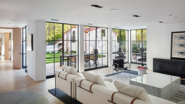 Bright open-plan contemporary living room with a large cream sectional sofa, black-framed floor-to-ceiling glazing, a white marble-look coffee table, black media console, and light timber flooring. The room opens visually to a landscaped outdoor area and is illuminated by abundant natural light.