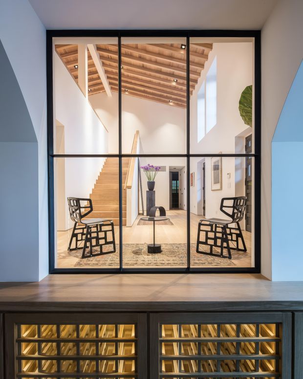 A view through a black-framed internal glass partition into a bright, double-height entry and stair hall. The space features a pale timber open-riser staircase, exposed timber ceiling rafters, white walls, black metal detailing, sculptural black lounge chairs, a central round table, and a patterned rug. Dark built-in wine storage with illuminated timber racks occupies the foreground below the glazing.