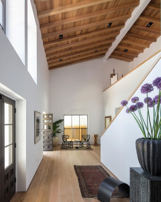 A bright, double-height residential entry hall with pale timber flooring, white walls, and a dramatic exposed timber rafter ceiling. A black-framed entry door is visible at left, while a sculptural staircase with a white balustrade and timber handrail rises at right. The space includes a small seating area near glazed doors, a console with a large black planter and purple allium-like flowers, and a patterned runner. Natural light enters through tall openings and glazing.