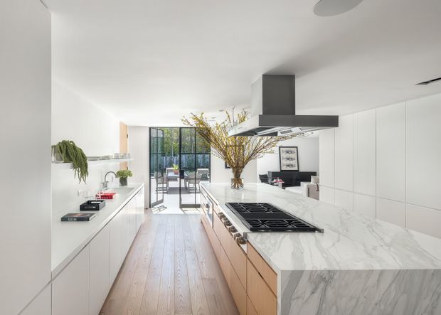 Bright contemporary open-plan kitchen with a long marble-look island and integrated gas cooktop, white full-height cabinetry, pale timber flooring, and a large stainless-steel rangehood. Glazed doors at the rear open toward a dining or outdoor area, with a living area visible beyond. The space has clean lines, abundant natural light, and restrained neutral styling.