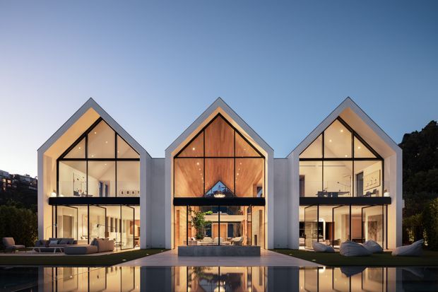 Contemporary multi-gabled residence viewed from across a reflecting pool at dusk. Three tall, sharply pitched glazed bays form a rhythmic facade, with dark slender window frames, warm timber-lined interiors, pale rendered or concrete-like exterior walls, and illuminated living spaces. Outdoor seating and a terrace occupy the foreground, while the pool mirrors the building.
