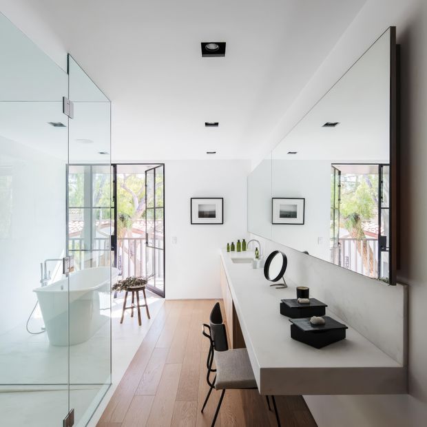Bright contemporary bathroom with a long floating vanity, integrated basin, wall-to-wall mirror, black countertop accessories, and two stools. A freestanding bathtub is enclosed behind frameless glass at the left, with full-height glazing opening to an outdoor balcony or garden view. The room has white walls and ceiling, recessed black downlights, warm timber flooring, and strong natural light.