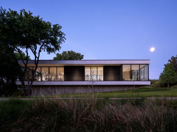 A modern two-storey rectangular residence photographed at dusk, with a flat roof, extensive floor-to-ceiling glazing, dark vertical screening panels, and a projecting upper-level terrace or balcony with glass balustrades. The home sits within a landscaped suburban or semi-rural setting with mature trees and grasses, while the illuminated interior is visible through the glazing.