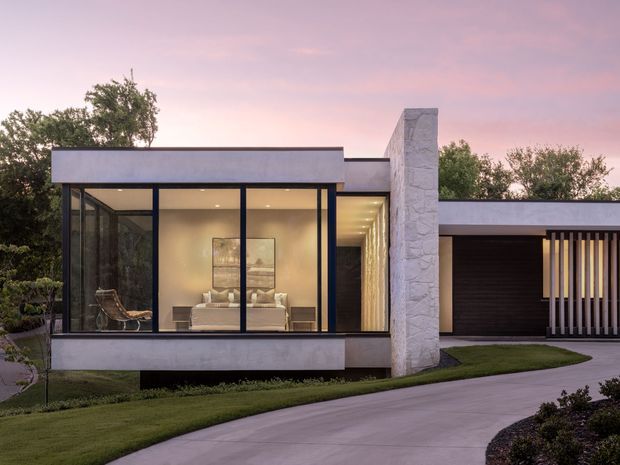 A dusk exterior view of a low-slung contemporary residence with broad floor-to-ceiling glazing, a projecting glass-fronted living area, pale rendered walls, a tall light stone-clad feature wall, and a flat-roofed garage or carport volume. A curved concrete driveway and landscaped lawn lead toward the house, while warm interior lighting creates a calm, refined atmosphere.