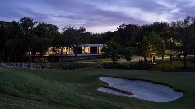 Twilight exterior view of a low, elongated modern building set within a landscaped golf-course-like grounds. The building has extensive warm-lit glazing, dark horizontal roof and wall lines, mature trees surrounding it, and a sculpted sand bunker in the foreground.