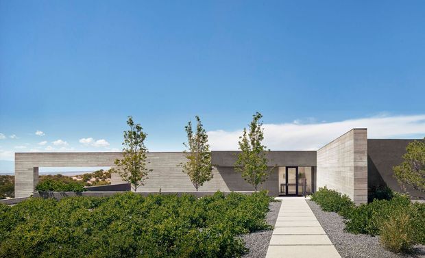 Low-slung contemporary residential building viewed from the landscaped front approach, with a minimalist horizontal composition, pale textured masonry walls, dark roof planes, deep rectangular openings, and a simple concrete-paver walkway through clipped planting. Clear blue sky and distant landscape are visible beyond the structure.
