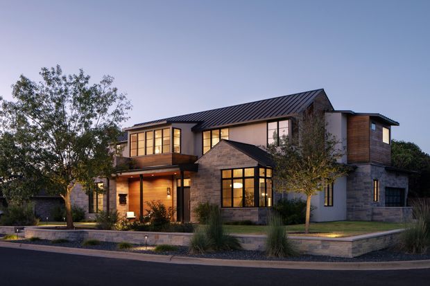 A two-storey contemporary residential house photographed at dusk. The home combines dark standing-seam metal pitched roofing, warm timber cladding, pale masonry walls, extensive black-framed glazing, and a projecting front entry. Low landscaped garden beds, ornamental grasses, a paved path, and a street frontage frame the facade.