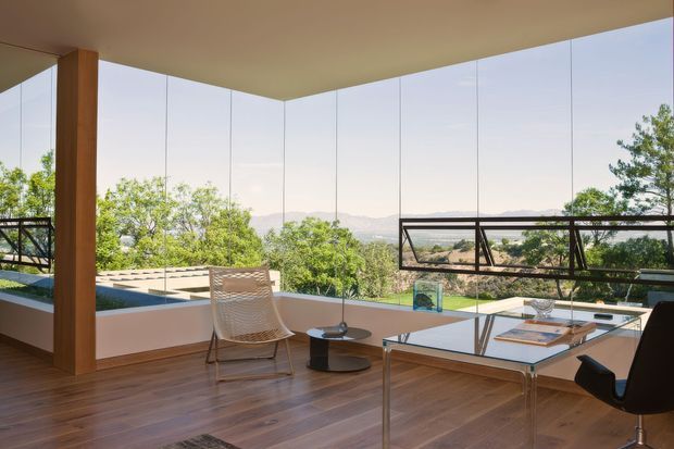Bright, modern glass-walled living or workspace overlooking a leafy landscape and distant hills. The room has warm timber flooring, a large timber structural column, floor-to-ceiling glazing, a glass-topped desk, contemporary chairs, and black-framed operable window panels. The open, elevated setting creates a spacious indoor-outdoor connection.