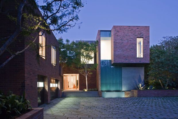 Twilight exterior view of a contemporary multi-level brick residence arranged around a paved courtyard. The composition features projecting brick volumes, large glazed openings, dark metal or glass wall panels, recessed illuminated windows, integrated planting beds, and broad cobblestone paving.