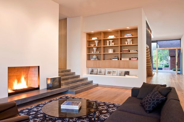 A warm, contemporary open-plan living room with a built-in brick fireplace, broad timber hearth, dark stone or concrete stair treads, and a large wall of custom timber shelving displaying books and ceramics. A dark sectional sofa and glass coffee table sit on a patterned rug over timber flooring. The room opens toward a bright glazed area and landscaped courtyard beyond.