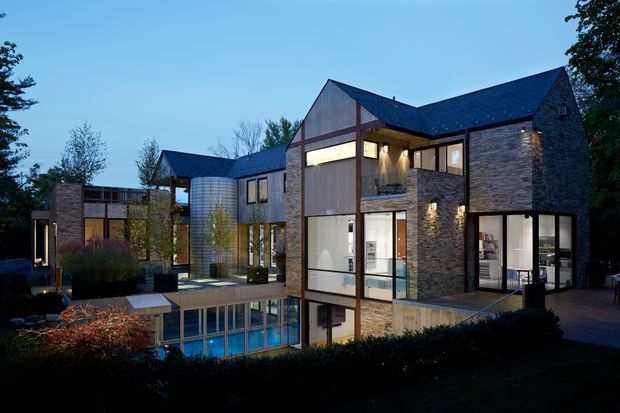 A contemporary multi-level residential building viewed from the landscaped exterior at dusk. The house has strongly articulated gabled roof forms, extensive floor-to-ceiling glazing, dark metal roofing, textured stone-clad walls, timber-toned vertical panels, and a partially enclosed courtyard or terrace with a visible blue-toned pool or water feature. Warm interior lighting contrasts with the cool evening exterior.