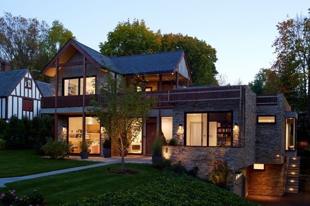 Exterior dusk view of a substantial multi-level residence set in landscaped grounds. The house combines dark brick and timber cladding, extensive glazing, projecting balconies, and steeply pitched gabled roofs with dark tiled roofing. A half-timbered neighbouring or connected wing is visible at left, while warm interior lighting creates an inviting glow through the windows. The site includes a lawn, paved walkway, planted garden beds, and illuminated exterior steps.