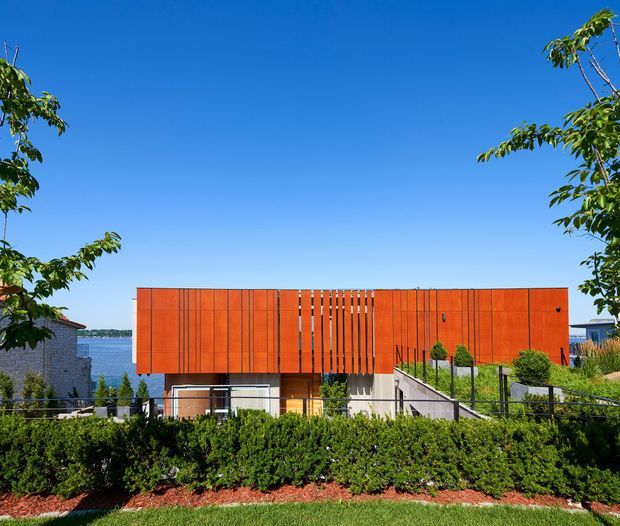 Exterior view of a contemporary multi-level residence set on a sloping site, with a vivid orange-red panelled facade and vertical screening elements. The building overlooks a body of water, with landscaped hedges, trees, a planted roof or terrace, and a stairway leading toward the house. Clear blue sky and strong sunlight create a bright, high-contrast setting.
