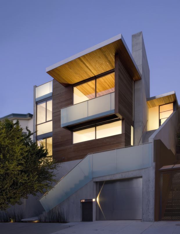 Twilight exterior view of a multi-level contemporary residence with strong geometric forms. The facade combines horizontal dark timber cladding, exposed concrete walls and fins, large glazed openings, translucent balcony panels, and warm timber-lined roof soffits. An external stair rises across the concrete lower level, with integrated warm lighting and a garage or entry zone at street level.